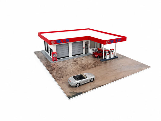 A miniature model of a gas station with a red and white canopy, gray garage doors, red fuel pumps, a small red car, and a silver convertible car on a printed ground surface, isolated on a white background.