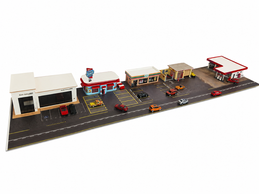 A miniature diorama model set depicting a retro roadside scene with multiple buildings, including a gas station, diner, and service station, along with small cars and trucks on a white background.