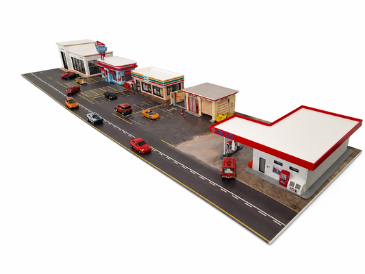 A miniature diorama model of a small town street scene with multiple buildings, including a gas station, convenience store, and service station, along with several toy cars on a gray road, shown against a solid white background.