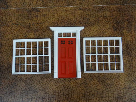 A 1:18 scale model of a house featuring a red door and white windows, placed on a textured background.