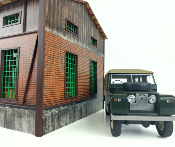 Model of a building with a brick facade and wooden roof next to a model of an old-fashioned green jeep.