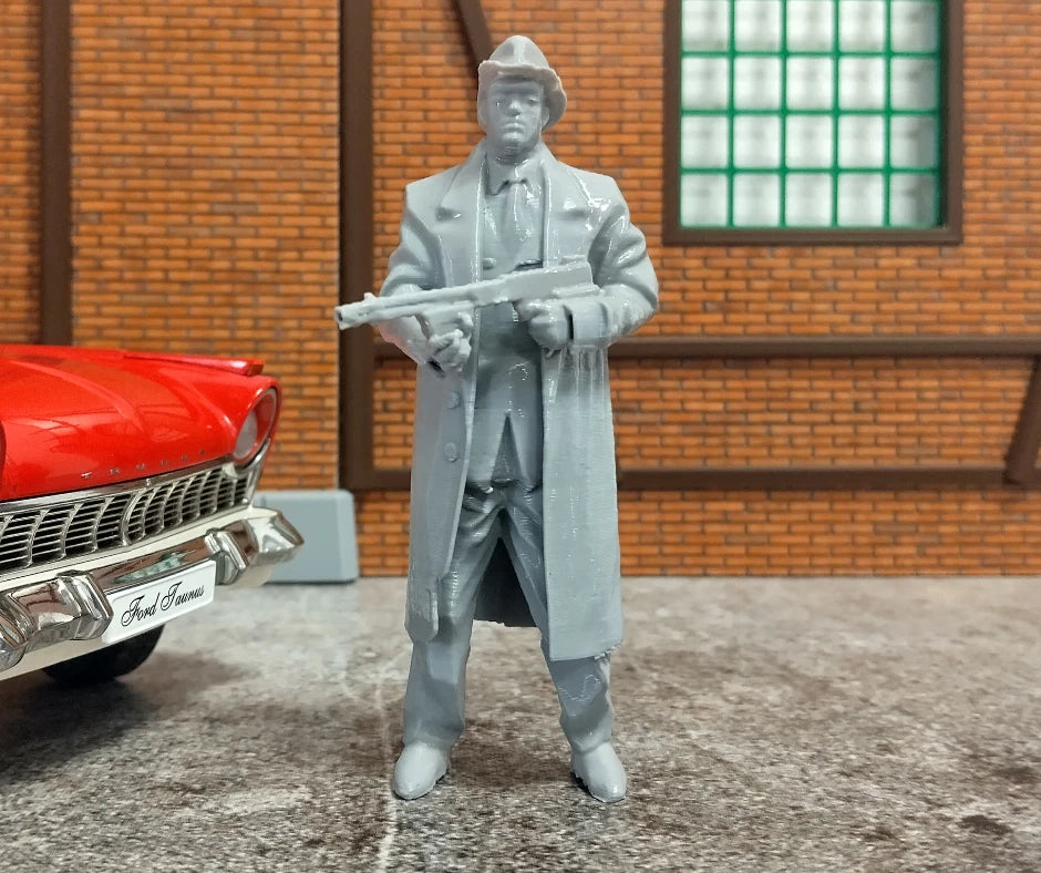 Model figure of a man in a suit holding a rifle next to a red car with a brick building in the background.