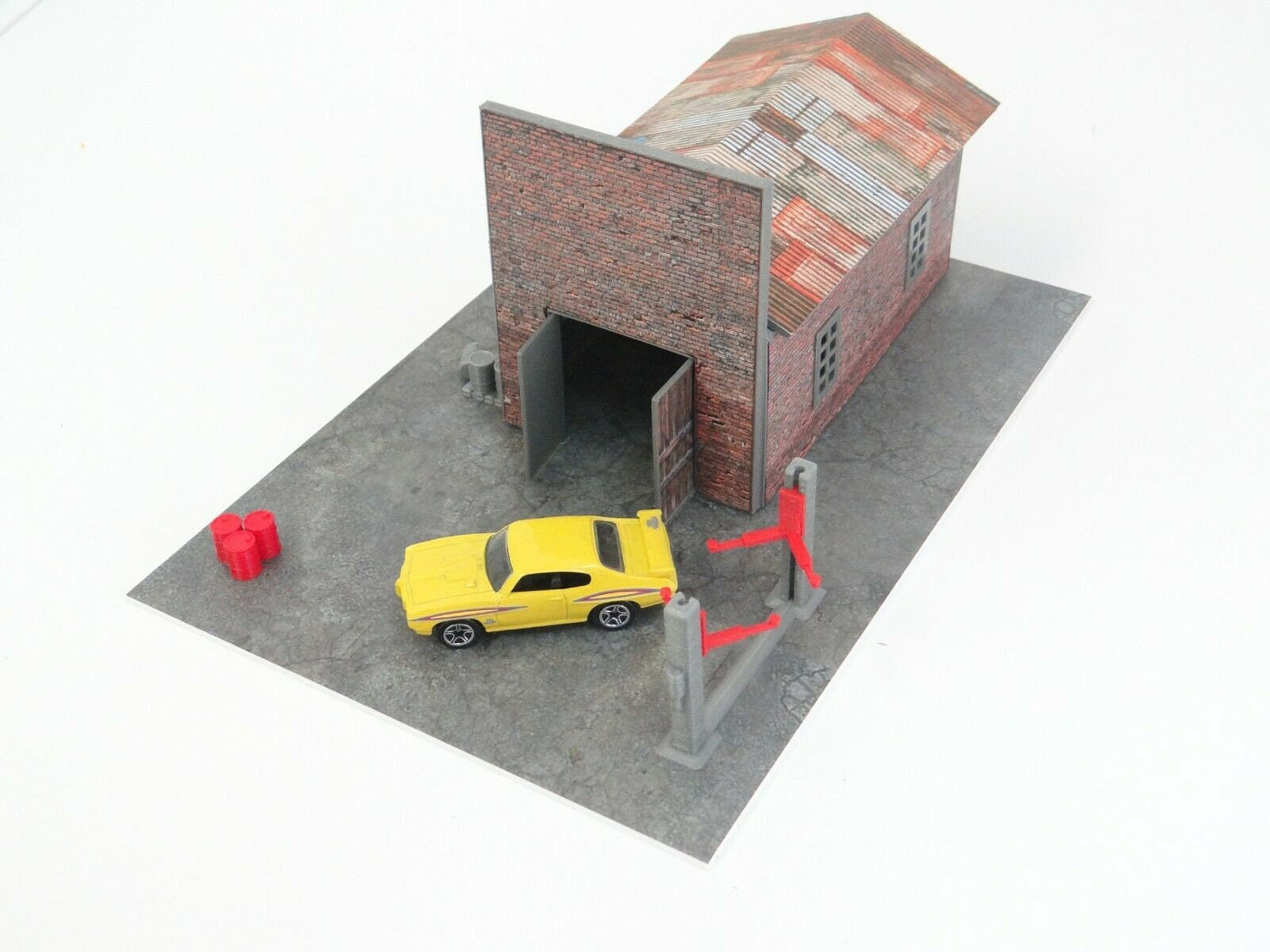 A diorama kit depicting a brick garage with a diecast car displayed inside. The kit includes various modeled elements like a parking base, car lifter, oil barrels, and a pallet.