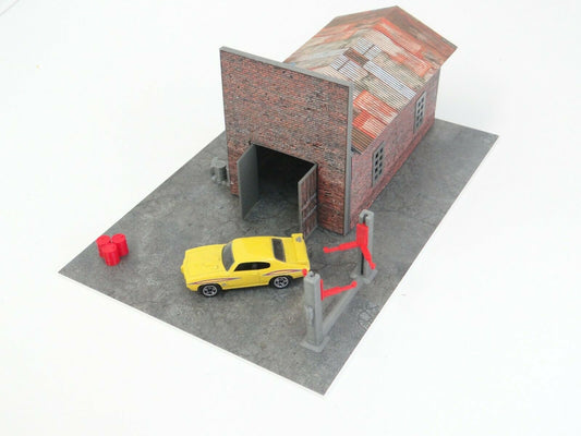 A diorama kit depicting a brick garage with a diecast car displayed inside. The kit includes various modeled elements like a parking base, car lifter, oil barrels, and a pallet.
